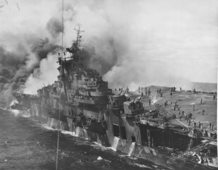 A fire and listing after a Japanese air attack on the Franklin, off the coast of Japan. Note fire hoses and crewmen on her forward flight deck and water streaming from her hangar deck. Photographed from USS Santa Fe.